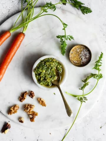 Carrot top pesto in a small bowl on a white surface, surrounded by walnuts and carrots