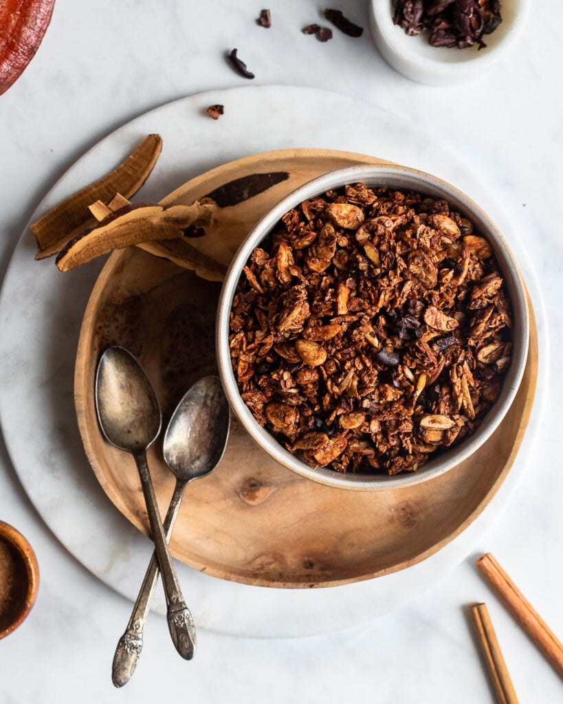 Dark chocolate granola in a bowl set on a wooden plate, garnished with cacao beans, reishi mushroom