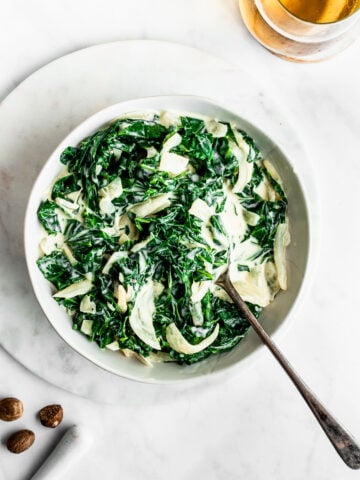 Creamed collard greens in a white bowl with a silver serving spoon on a marble background