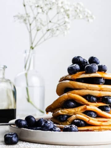 Stack of einkorn pancakes garnished with blueberries and drizzled with maple syrup on white plate.