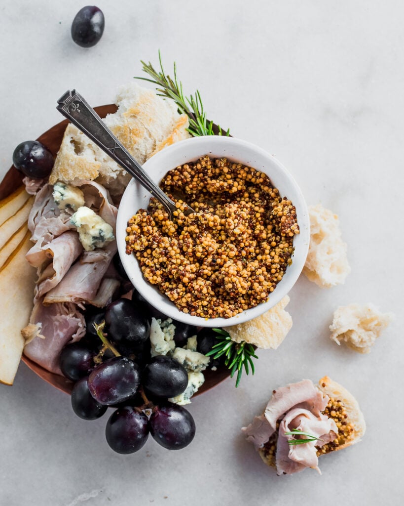 Fermented whole grain mustard in a bowl on a charcuterie platter with bread, grapes, ham, and cheese.