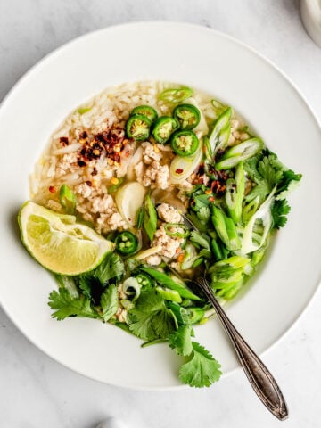 Ginger and chicken soup in a white bowl garnished with cilantro, chili pepper, scallions and lime