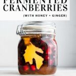 Pinterest pin featuring fresh cranberries and sliced fresh ginger in a honey-filled fido jar on a marble surface