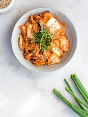Kimchi in a stoneware bowl on a marble background, garnished with sesame seeds and green onions.