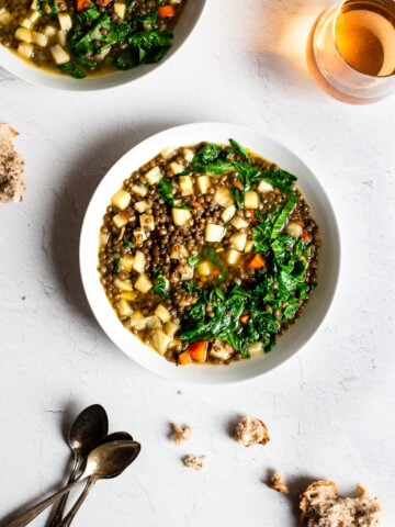 Two bowls of lentil stew on a white surface. Bread crumbs are on the table as well as a glass of beer.