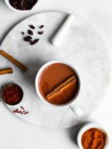 mayan hot chocolate in a white mug on a marble surface, garnished with cinnamon and annatto.