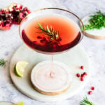 Pomegranate spritzer in a coupe glass garnished with pomegranate arils and rosemary next. Opened pomegranate, quartered lime, and rosemary branches in background.