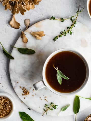Herbal sipping broth in a white mug, herbs and mushrooms in the background