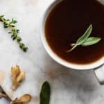 Herbal sipping broth in a white mug, herbs and mushrooms in the background
