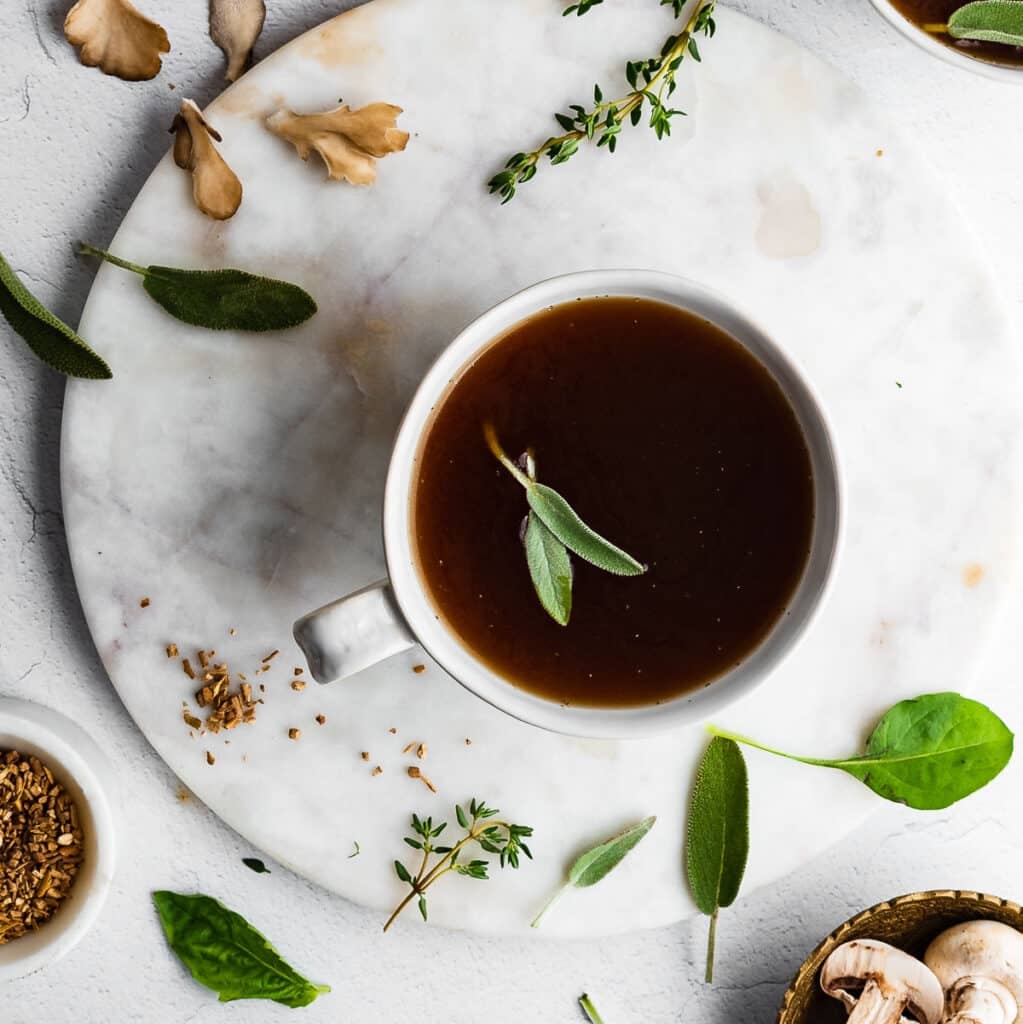 Herbal sipping broth in a white mug, herbs and mushrooms in the background
