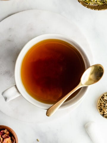 Herbal tea in a white mug with a golden spoon next to small bowls filled with passionflower and rose slightly out of frame