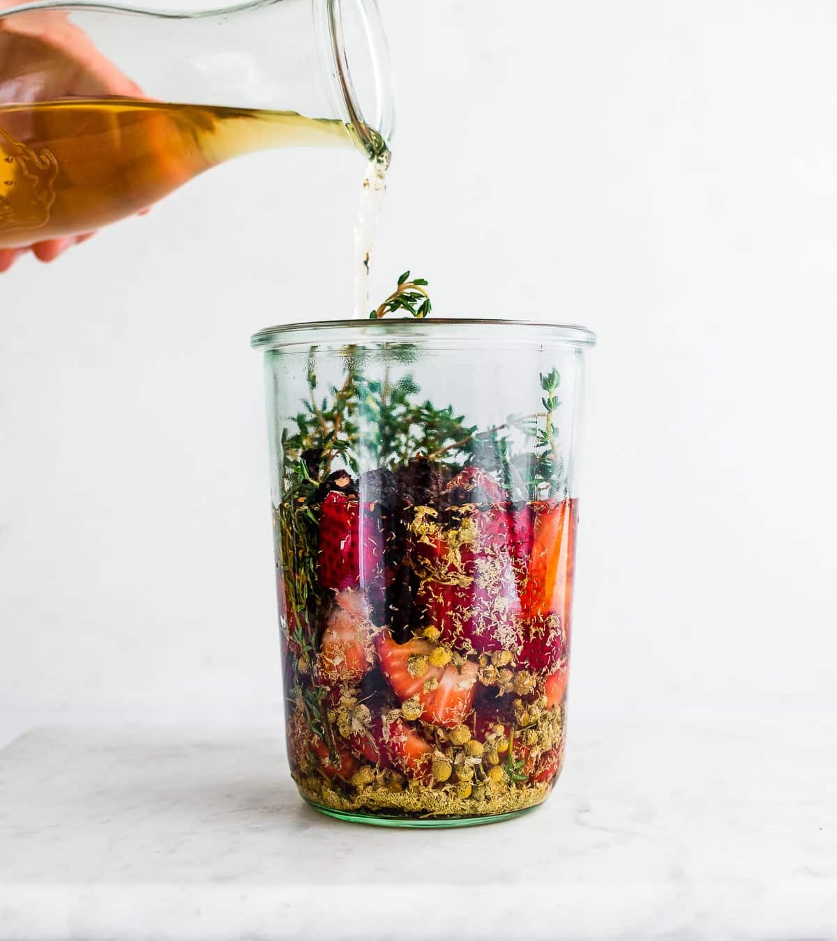 Pouring vinegar into a clear jar of strawberries and herbs to make a shrub.